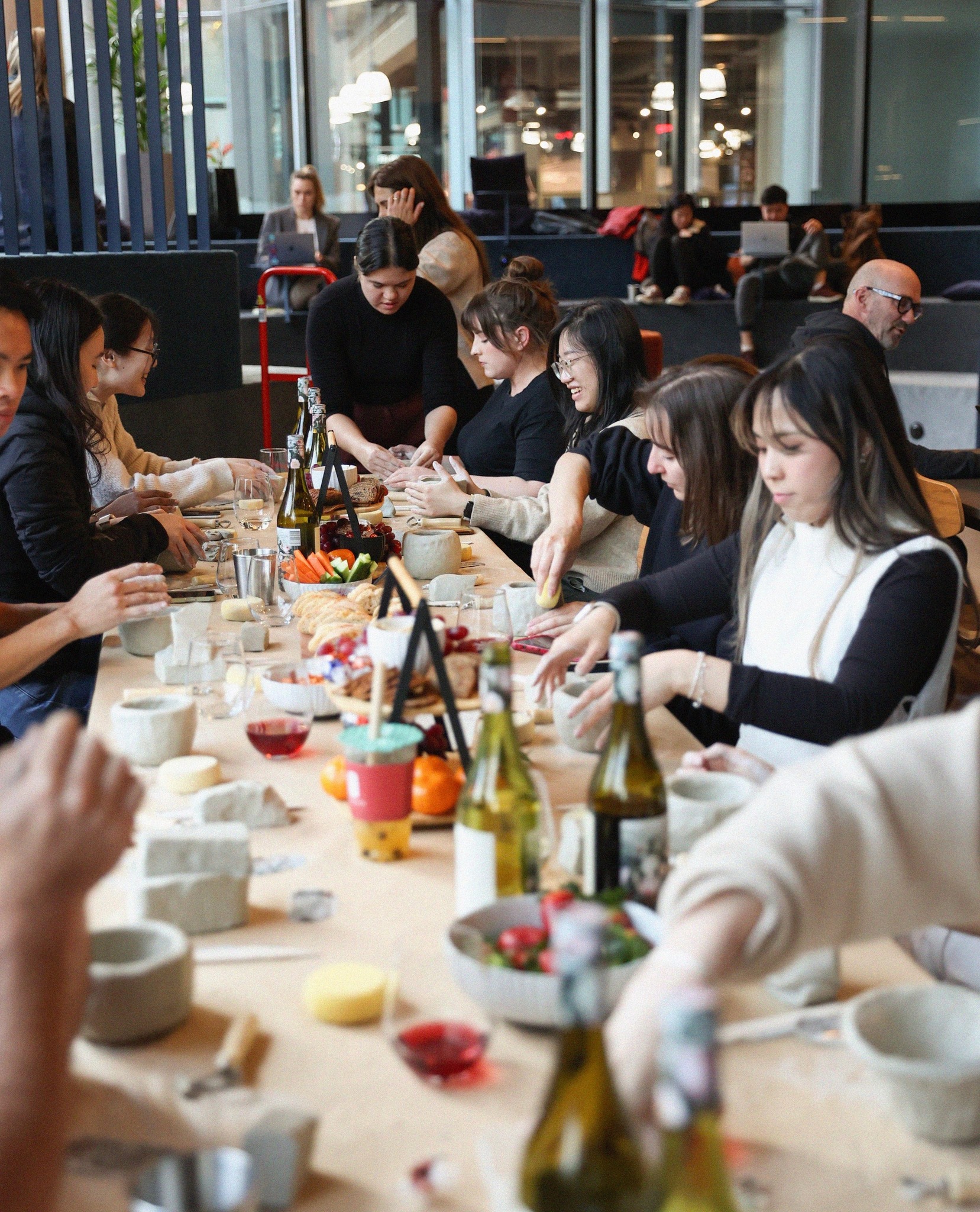 A group of people are gathered around a long table, seemingly preparing food and drinks in a well-lit, modern indoor space.