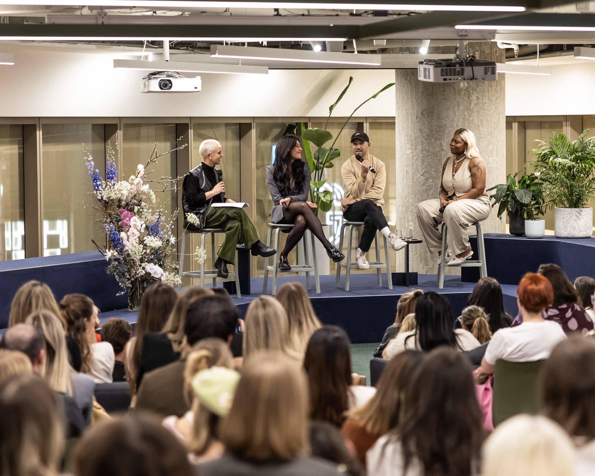 A panel of four people are sitting on stools on a stage in front of a large audience in a brightly lit indoor venue.