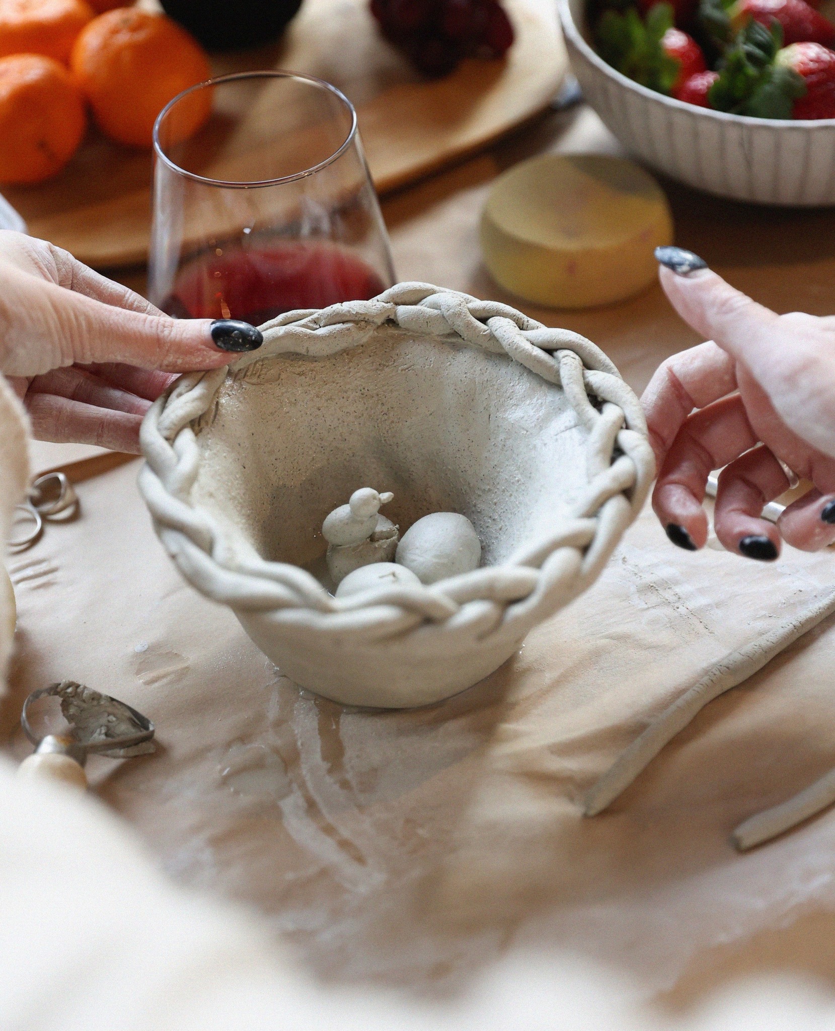 Hands crafting a clay bowl decorated with a braided rim, holding small clay figures, with oranges, strawberries, and a glass in background.