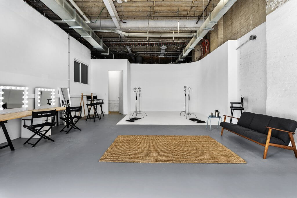 Wide shot of a bright photo studio featuring director's chairs at makeup stations, photography equipment, a tan rug, and a gray sofa.