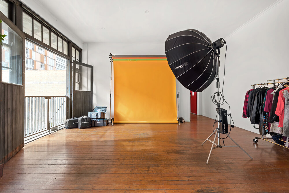 Empty photo studio features backdrop, lighting equipment, rolling rack with clothes, hardwood floor, and natural light from open doors.
