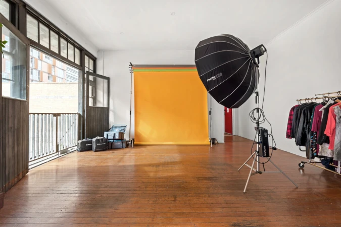 Empty photo studio features backdrop, lighting equipment, rolling rack with clothes, hardwood floor, and natural light from open doors.