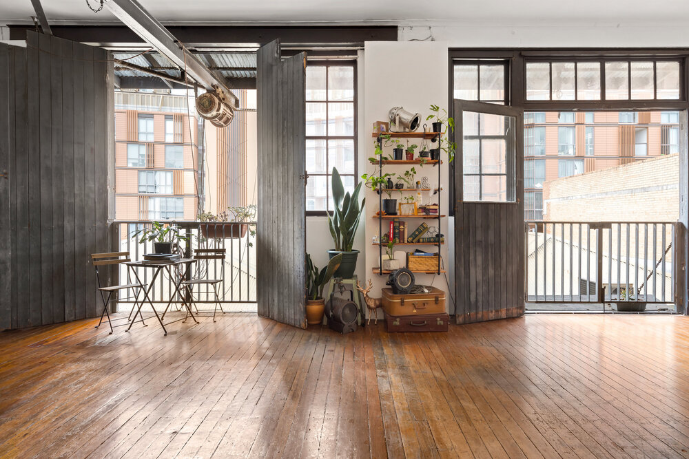 Spacious studio apartment interior with wooden floor, large windows, houseplants, folding doors, and balcony overlooking a city building.