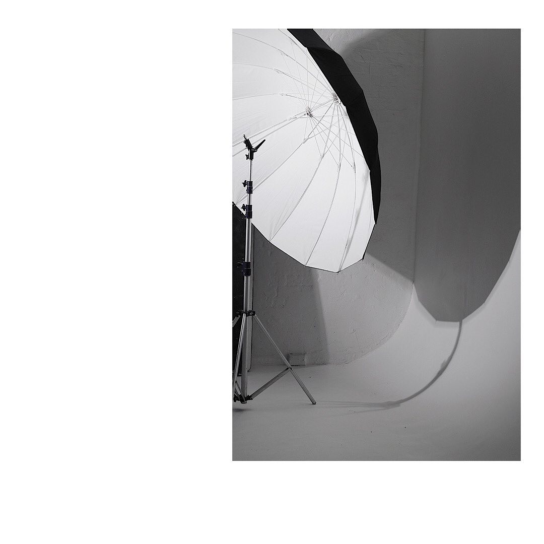 Monochromatic photo studio setup featuring a large white umbrella reflector on a stand, casting soft shadows on a textured white wall.