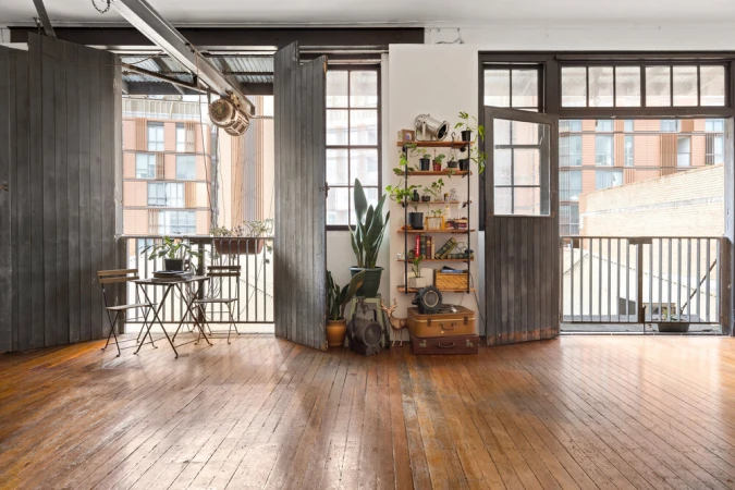 Spacious photo studio with polished wooden floors, large dark wooden folding doors, open balcony, and shelving units filled with plants.