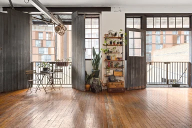 Spacious photo studio with polished wooden floors, large dark wooden folding doors, open balcony, and shelving units filled with plants.