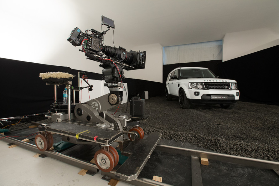 Automotive studio shoot features a white Land Rover Discovery with a camera and dolly on a gravel surface against a black and white backdrop.
