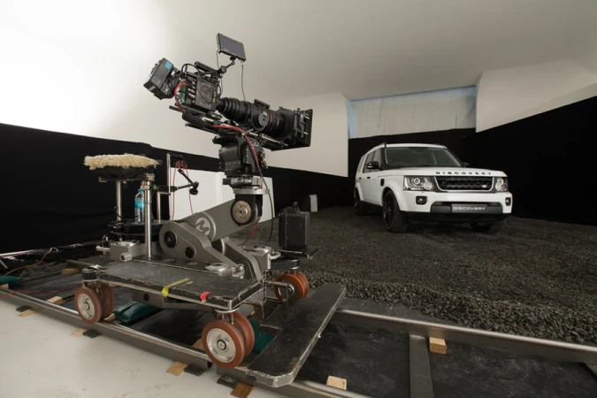 Automotive studio shoot features a white Land Rover Discovery with a camera and dolly on a gravel surface against a black and white backdrop.