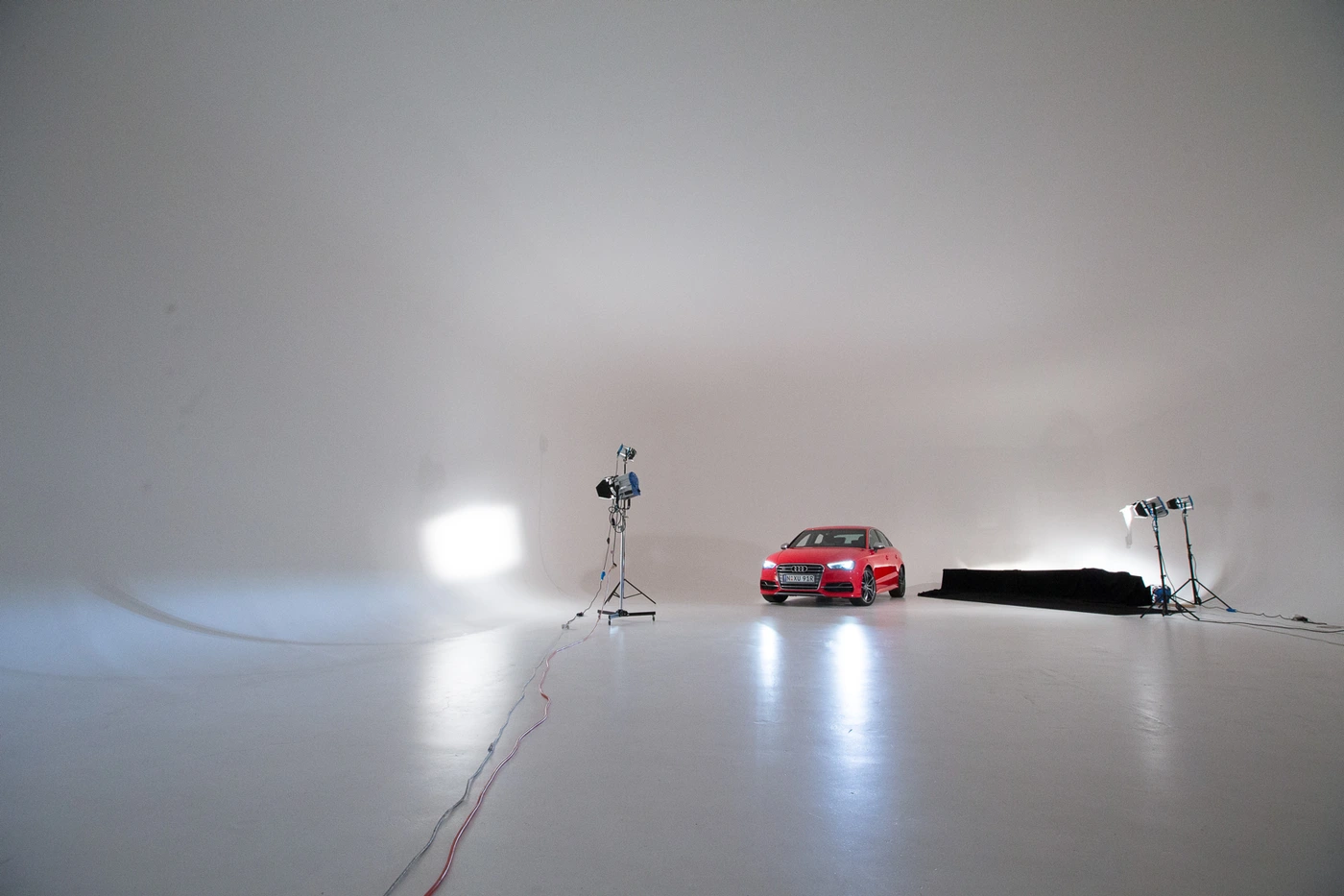 Red car parked inside a large white photo studio with studio lights and cables