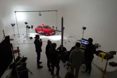 A crew of people standing in a large white studio watching a red car parked under overhead lighting equipment.