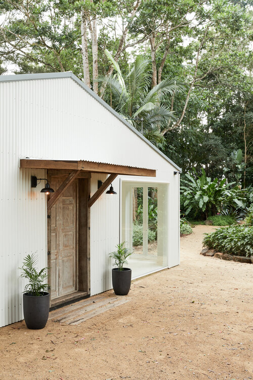 Exterior of a white corrugated metal building with a wooden door and awning, framed by lush greenery and potted plants.
