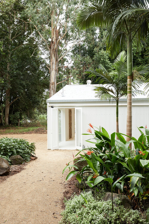 Exterior of a white building with a metal roof, set amidst lush greenery and tropical plants, showcasing architectural design and landscape.
