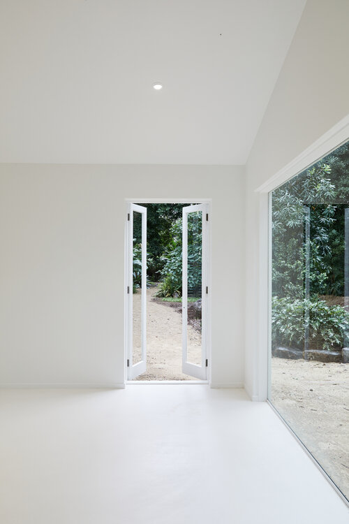 Bright, minimalist interior with white walls and floors, featuring open double doors leading to a lush green garden and a large window.