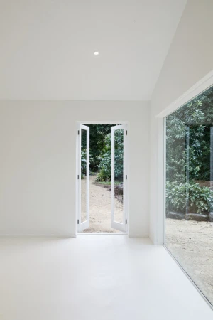 Minimalist white interior room featuring open double glass doors looking out onto a lush green garden path.