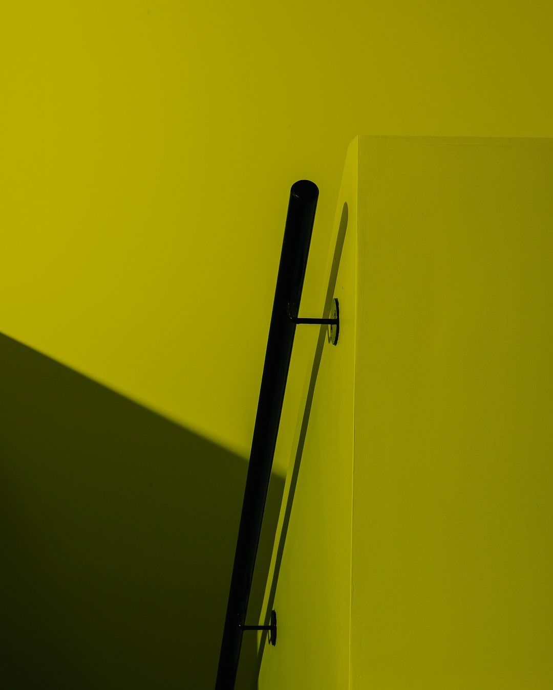 Minimalist architectural shot of a black handrail mounted on a chartreuse wall, creating geometric shapes and a strong interplay of light and shadow.