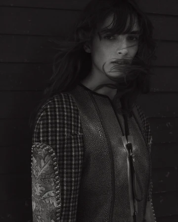 Portrait of a woman with dark hair blowing across her face, wearing a mixed-pattern jacket against a dark wood wall.
