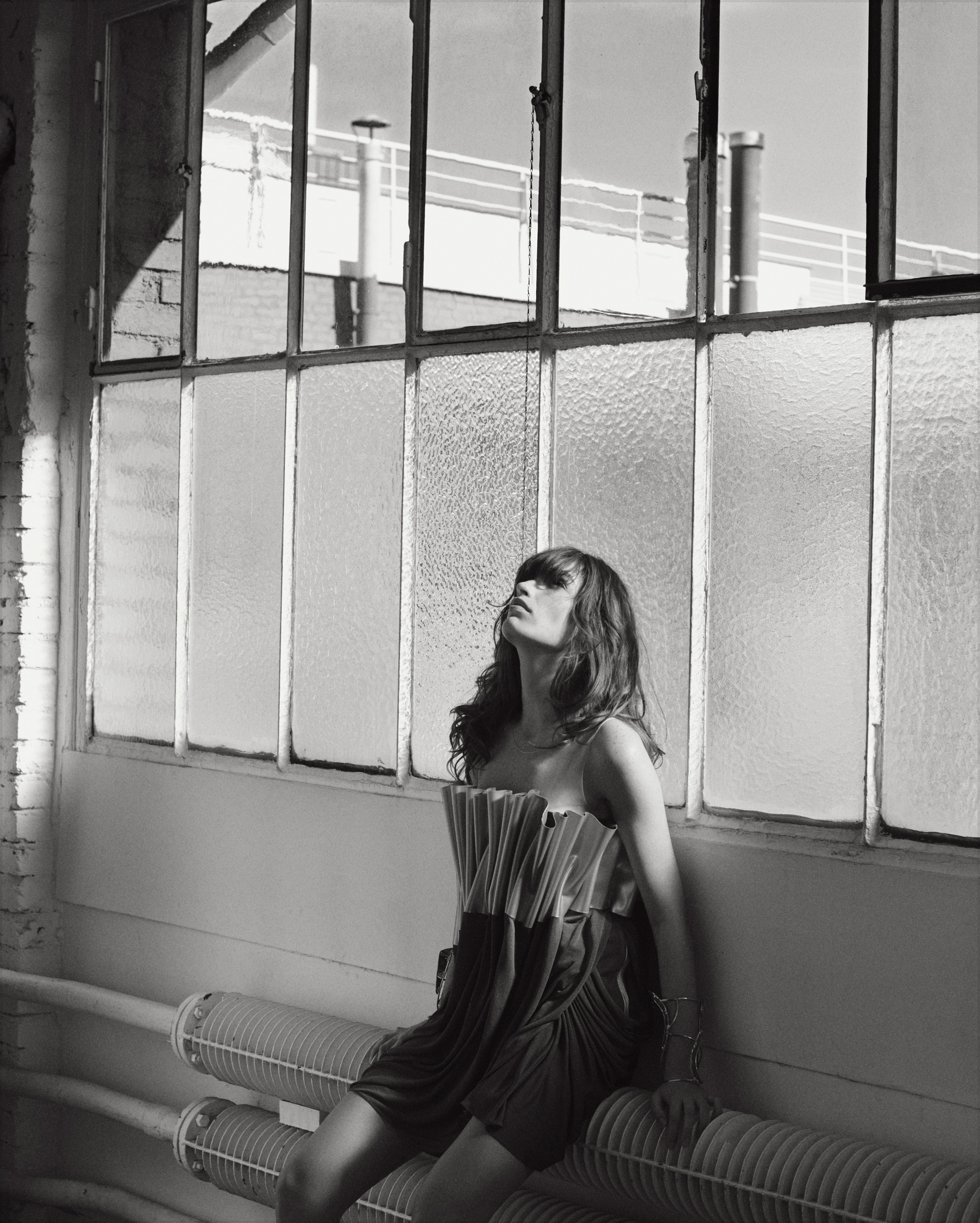 Monochrome portrait of a woman in a pleated strapless dress seated against industrial warehouse windows by Eminente Creative Production