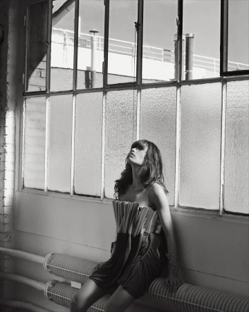 Monochrome portrait of a woman in a pleated strapless dress seated against industrial warehouse windows by Eminente Creative Production