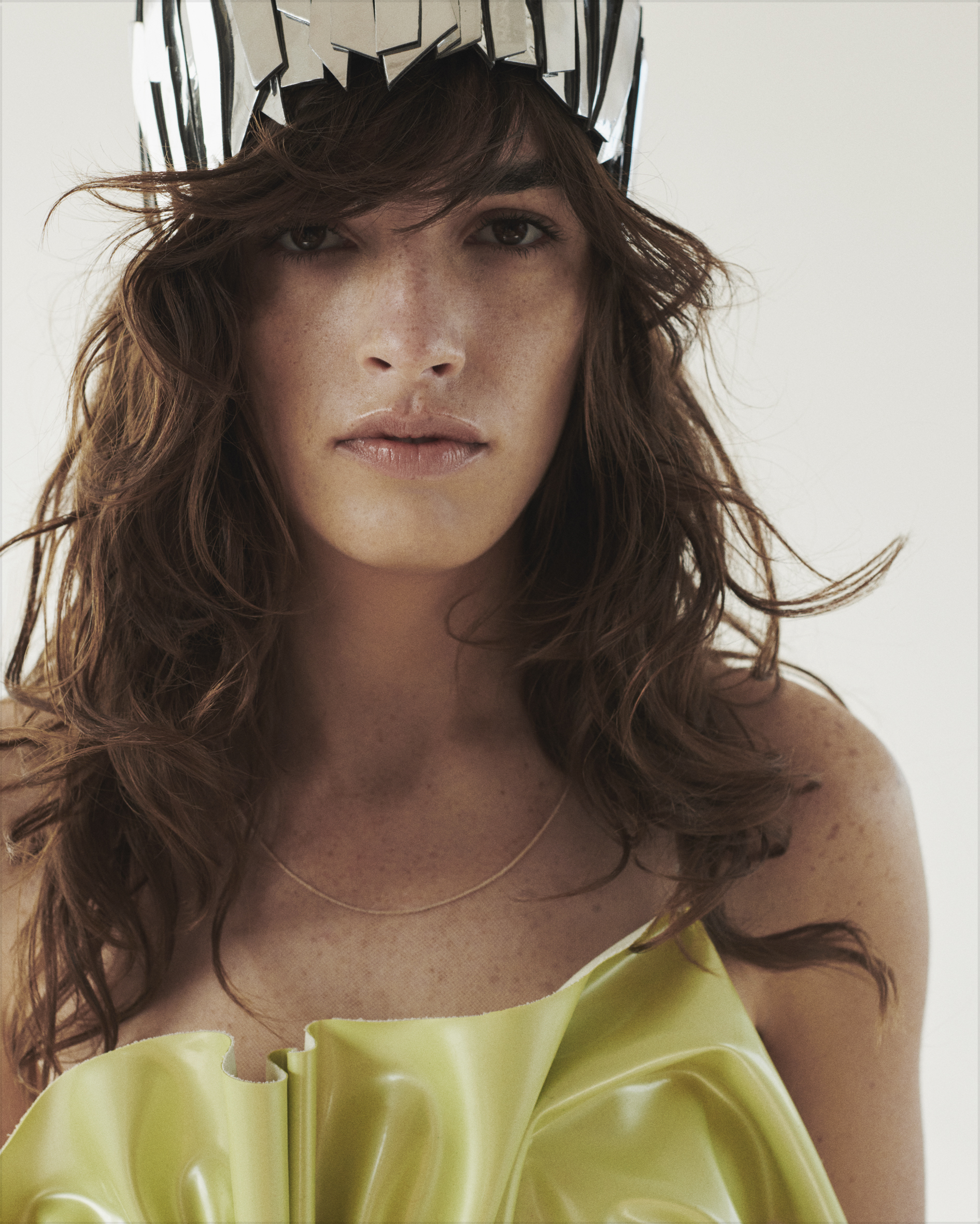 Close-up portrait of a person with wavy brown hair, a shiny green top, and a silver headpiece against a bright background in a studio.