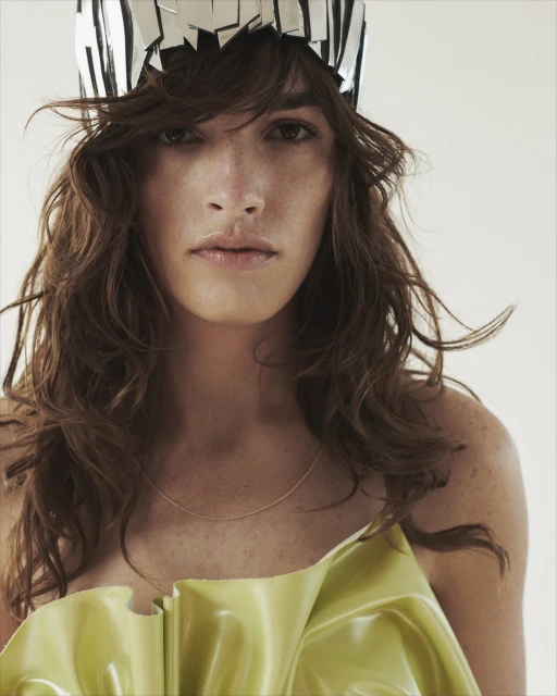 Close-up portrait of a person with wavy brown hair, a shiny green top, and a silver headpiece against a bright background in a studio.