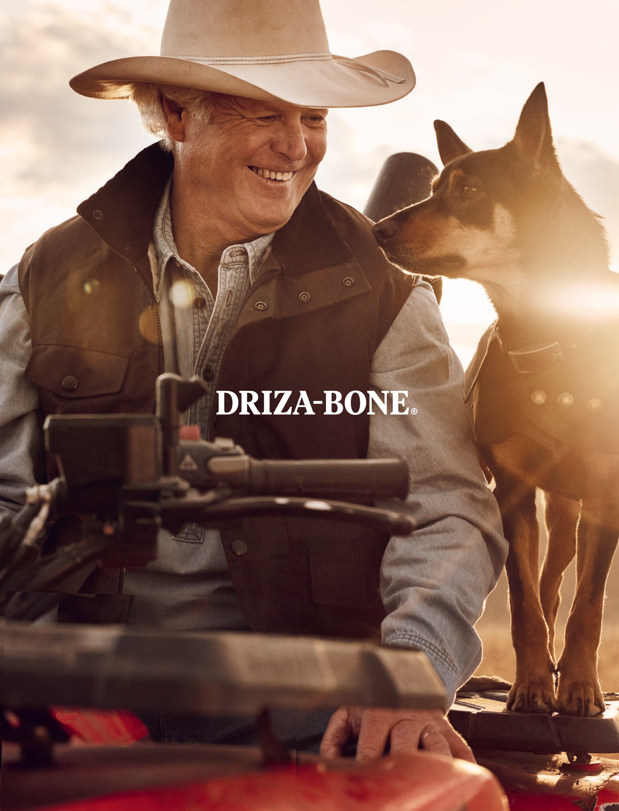 An adult male wearing a cowboy hat smiles while posing next to a dog in an outdoor environment with warm, natural lighting.