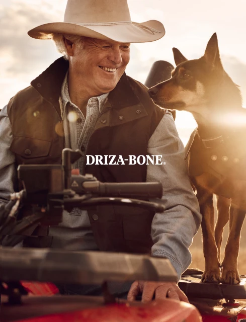 An adult male wearing a cowboy hat smiles while posing next to a dog in an outdoor environment with warm, natural lighting.