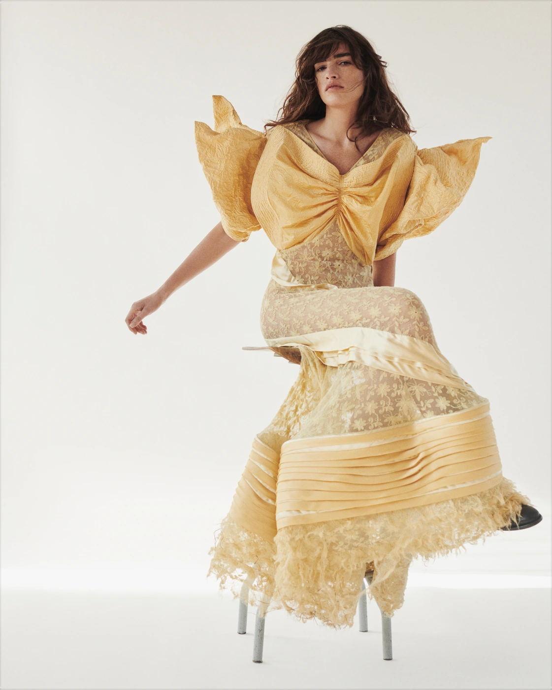 Woman in a textured yellow gown with sculptural puffed sleeves seated on a metal stool against a white backdrop.