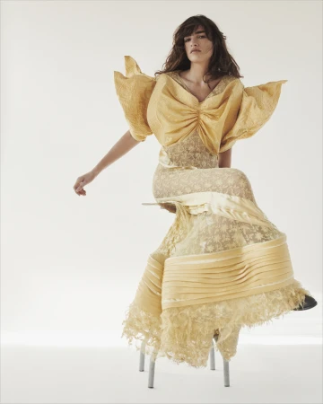 Woman in a textured yellow gown with sculptural puffed sleeves seated on a metal stool against a white backdrop.