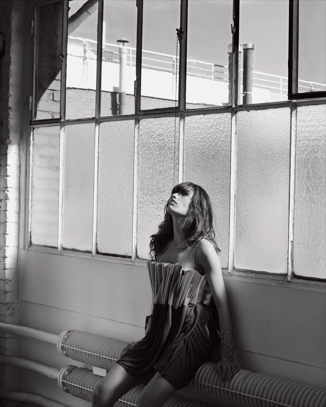 Monochrome portrait of a woman in a pleated strapless dress seated against industrial warehouse windows by Eminente Creative Production