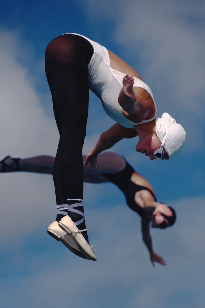 Two women in swimsuits and swim caps diving through the air against a clear blue sky background.