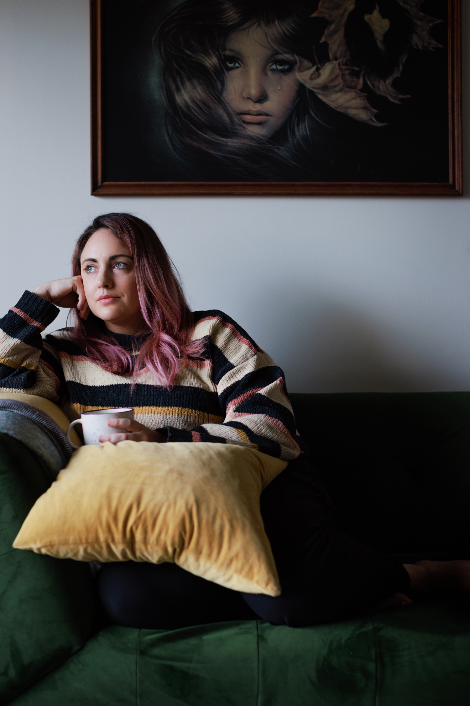 Adult female sits on green velvet couch with yellow pillow, holding white mug, in a room with art on the wall.