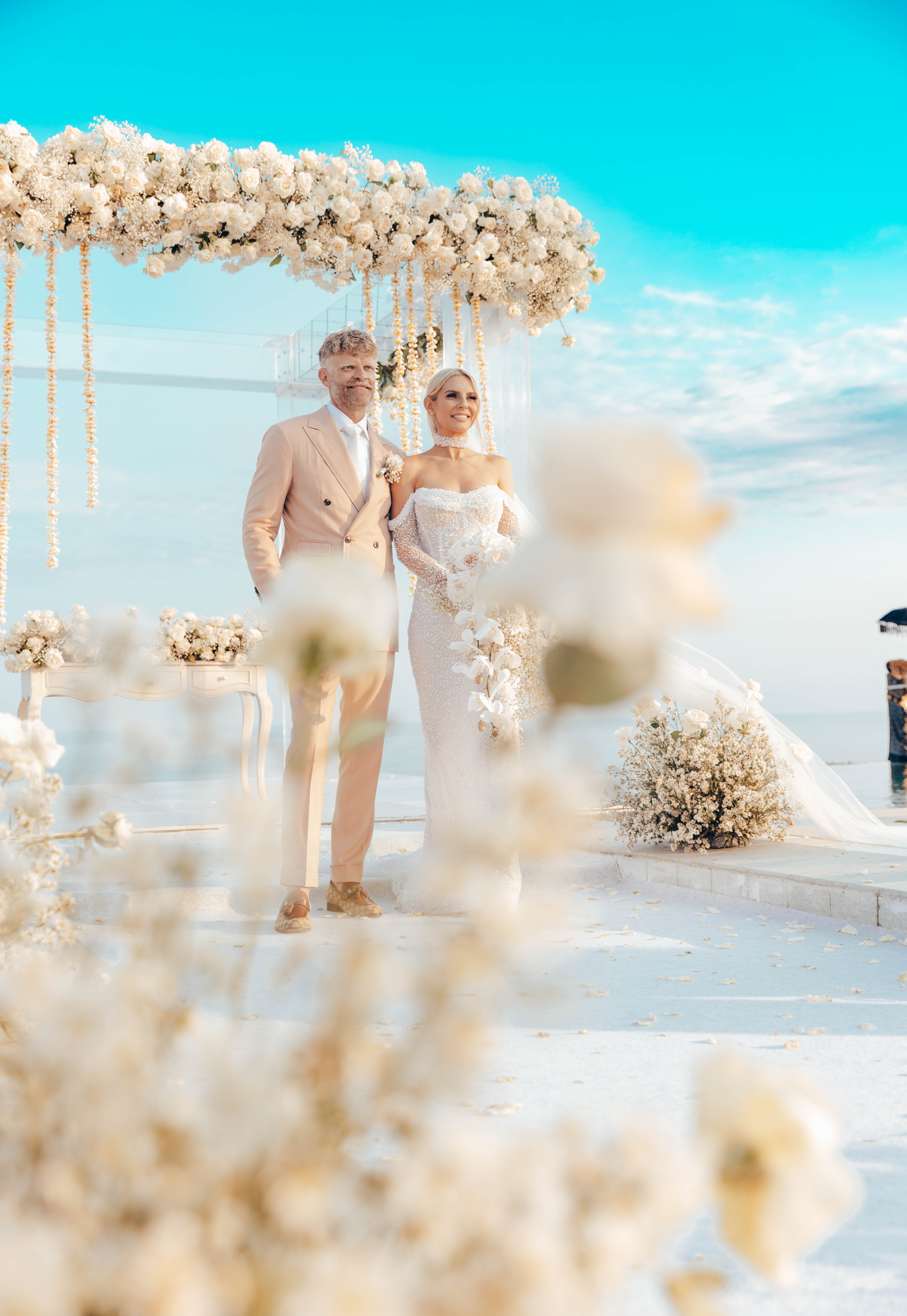 Couple in wedding attire stands under floral arch on sunny day. Bride wears elegant dress, groom in suit. Celebratory, romantic.