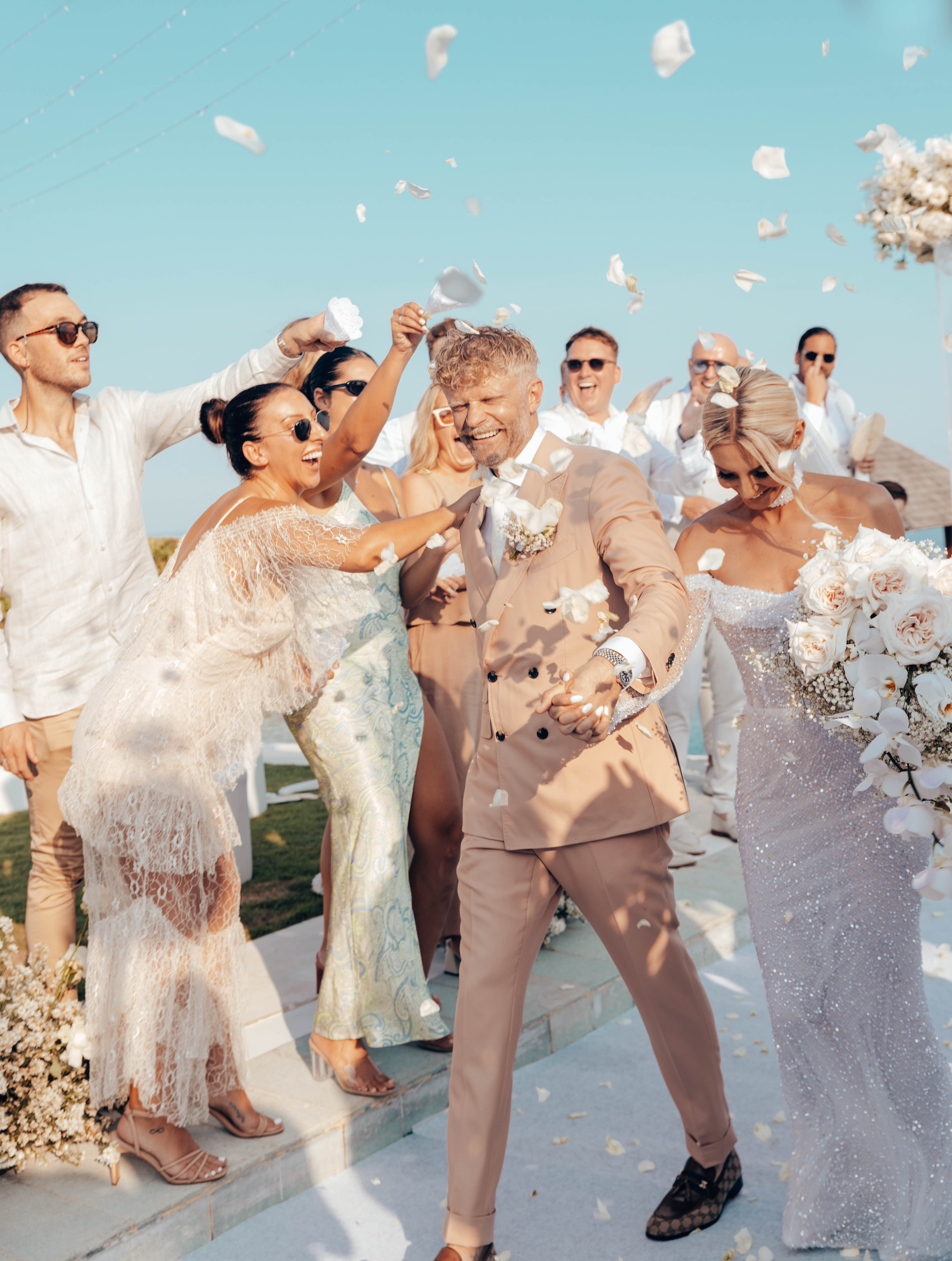 Wedding celebration: A happy couple walks down the aisle as guests toss flower petals under a clear blue sky in an outdoor setting.