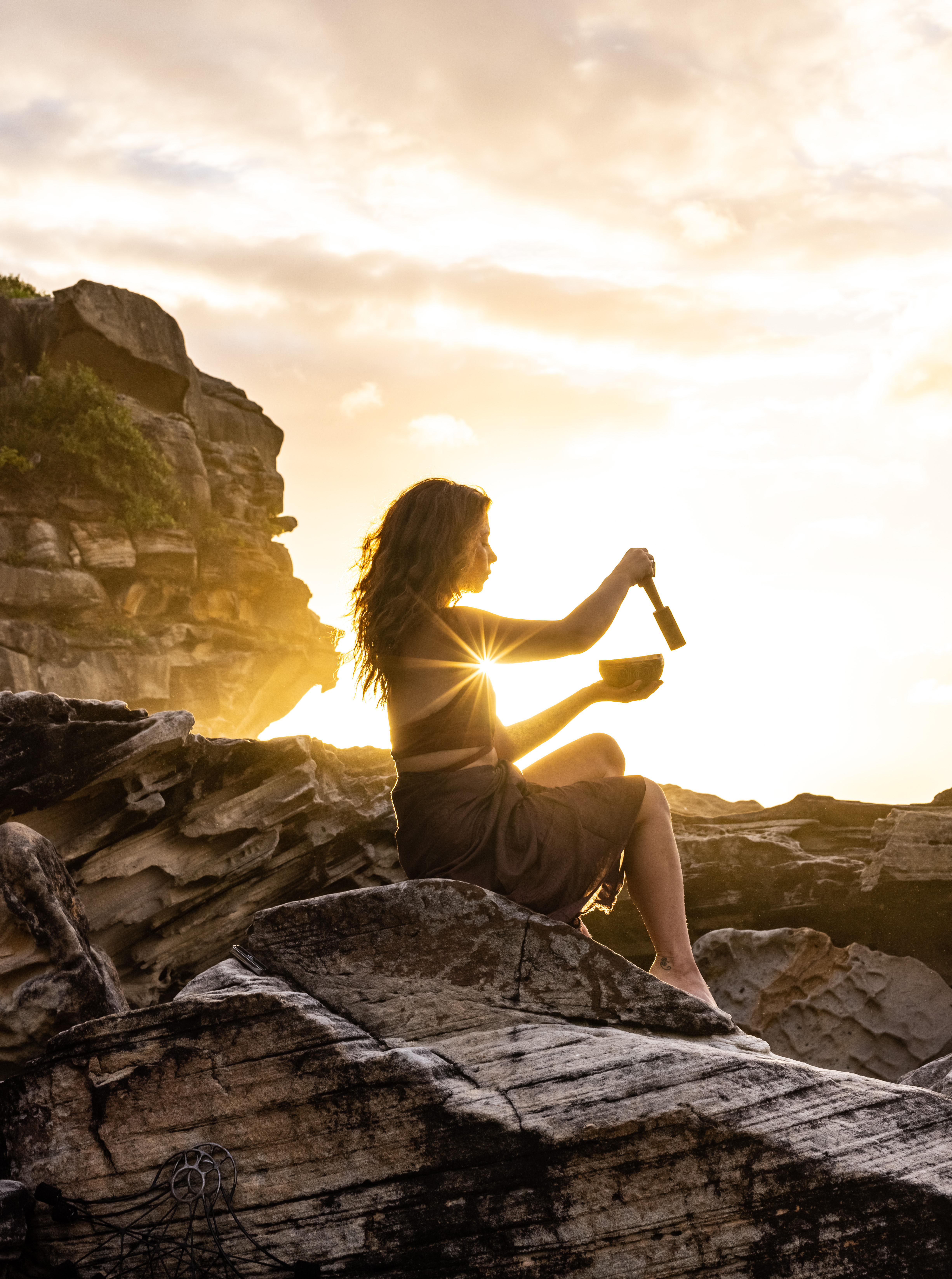 Adult female sits on rocky coast at sunset, playing singing bowl. Warm light on rocks, cliff, and ocean with cloudy golden sky.