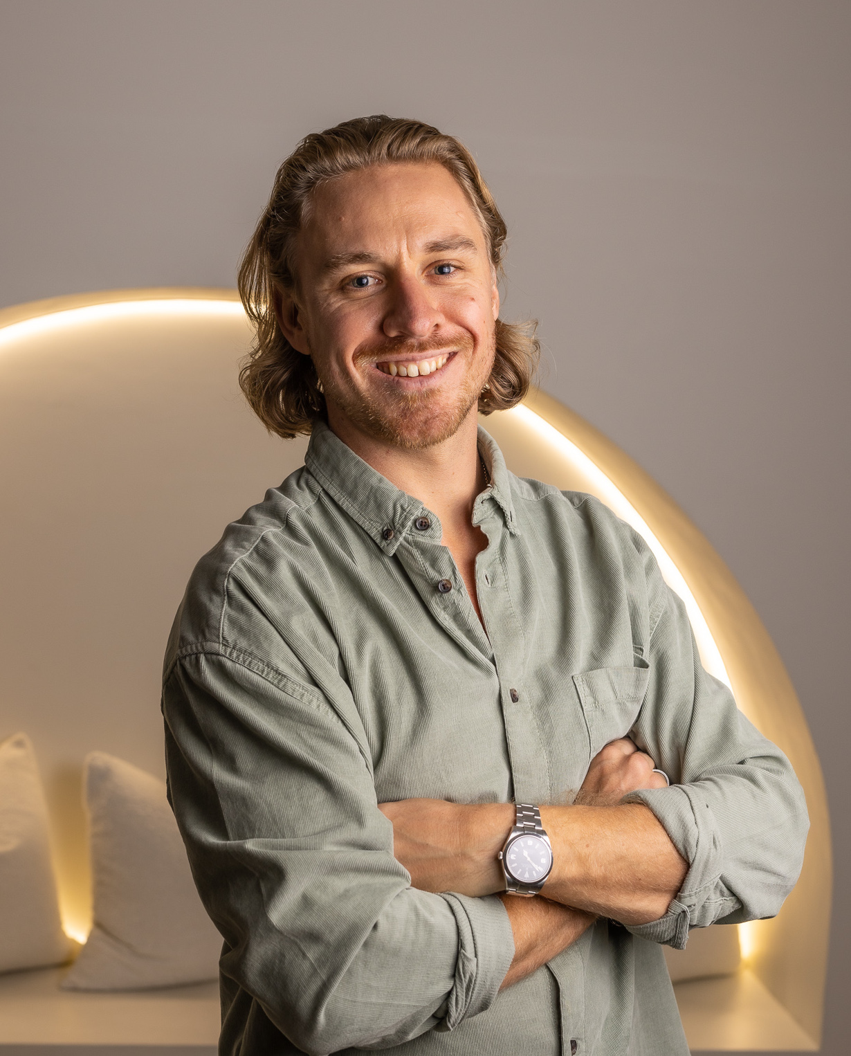 Smiling man with folded arms poses indoors wearing a green button-down shirt and a wristwatch, with soft studio lighting.