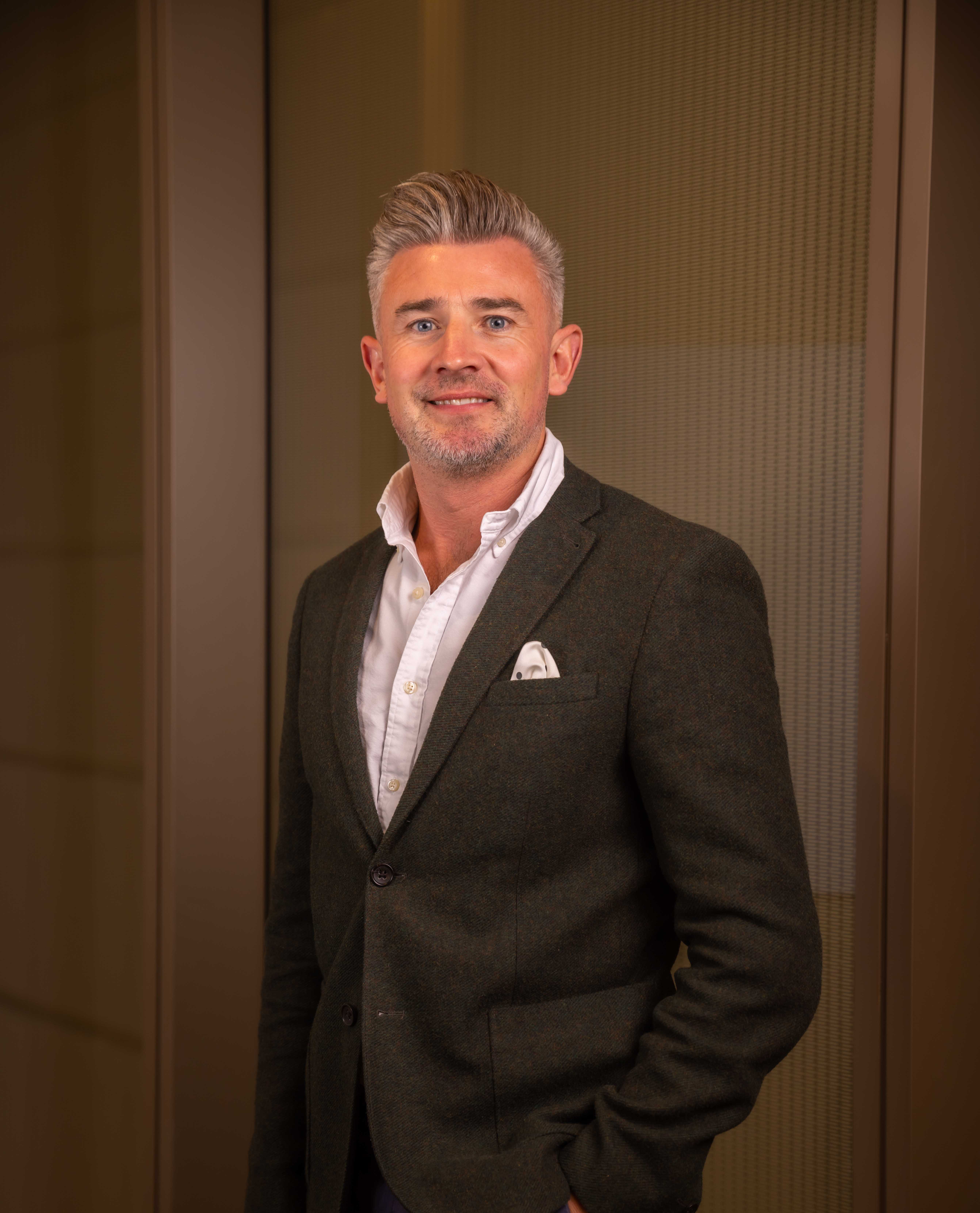 A stylish adult male in a blazer and white shirt poses indoors against a neutral wall, offering a friendly smile. Portrait orientation.