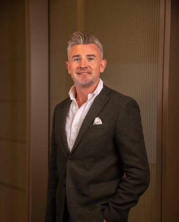 Portrait of a white man in his 40s wearing a dark grey blazer and a white shirt against an office background.