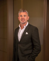 Portrait of a white man in his 40s wearing a dark grey blazer and a white shirt against an office background.