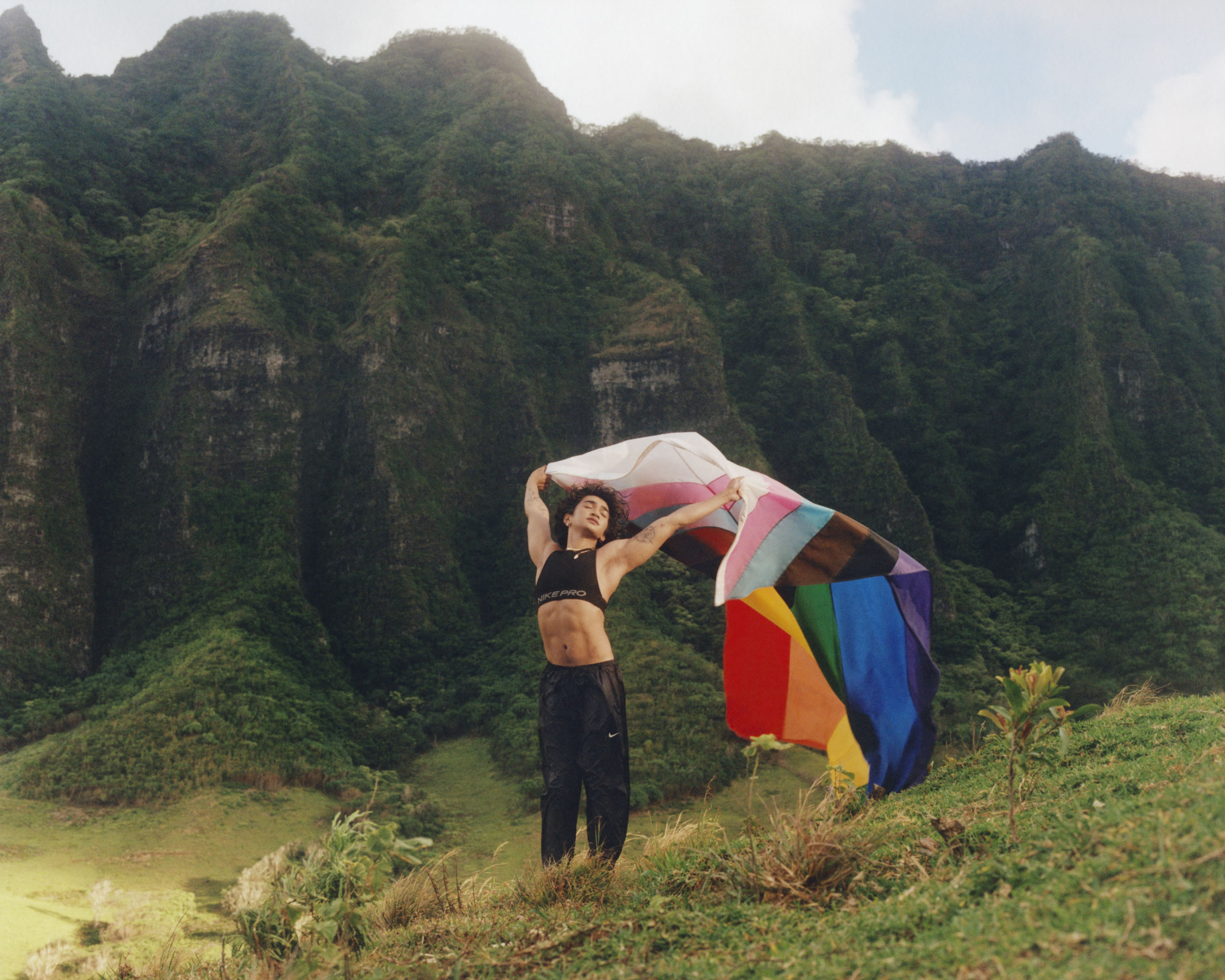 Athletic woman standing on a grassy hill with a colorful flag, posing with a mountain range in the background on a sunny day.