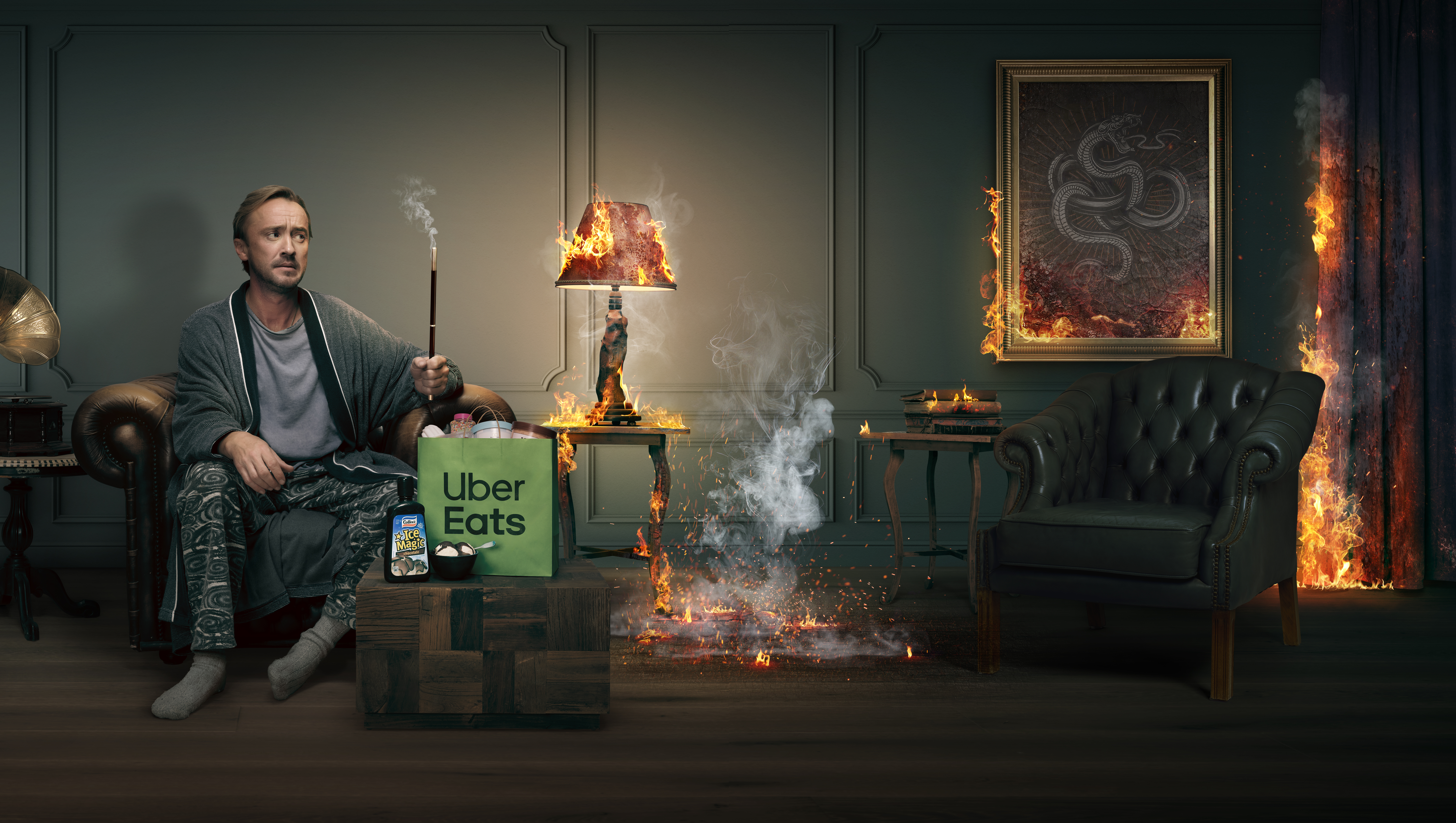 Tom Felton sitting on a leather sofa in a living room, surrounded by fire and holding a wand with Uber Eats bag nearby.