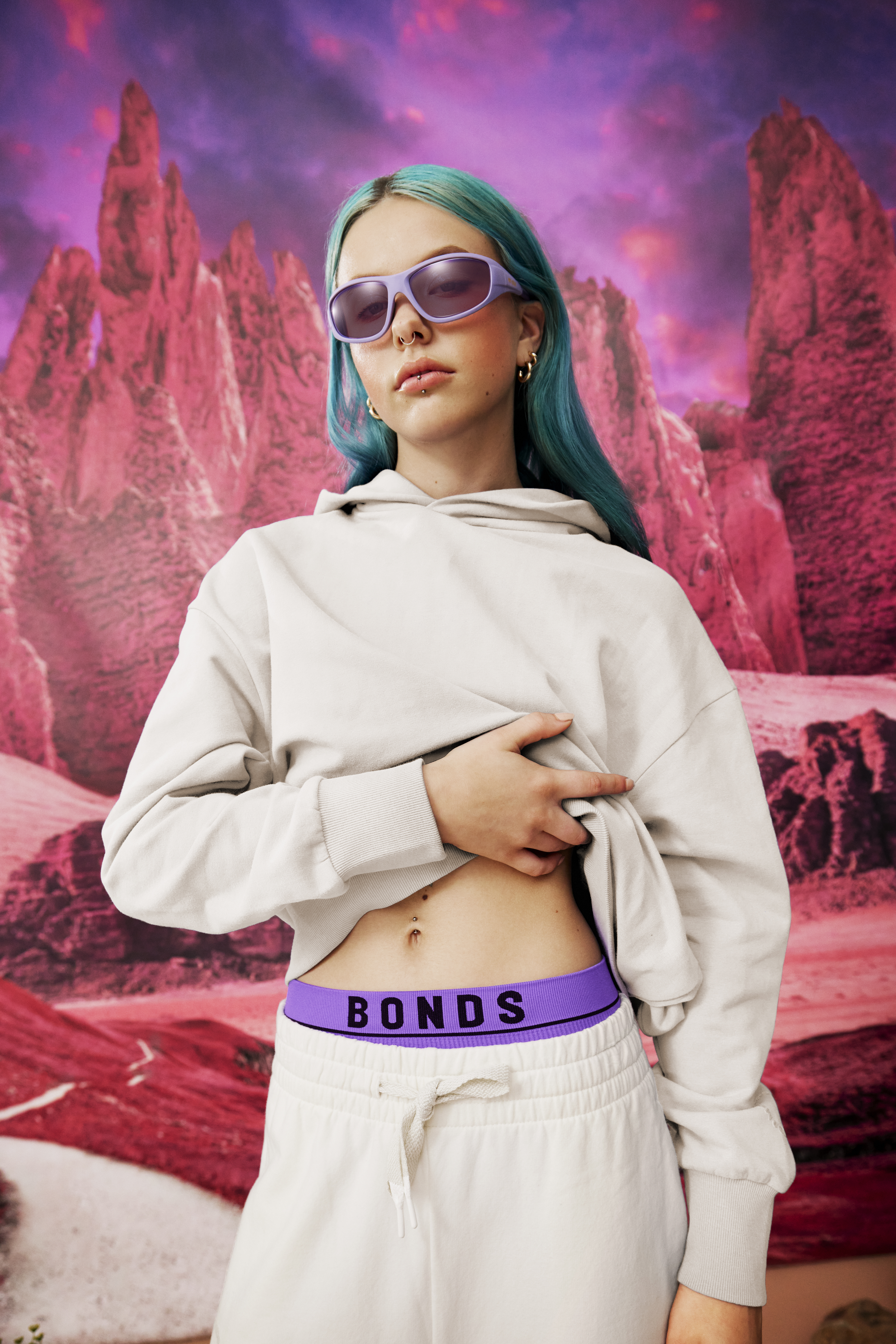 Young woman with blue hair poses in a beige hooded sweatshirt and sweatpants with purple sunglasses in front of a red landscape.