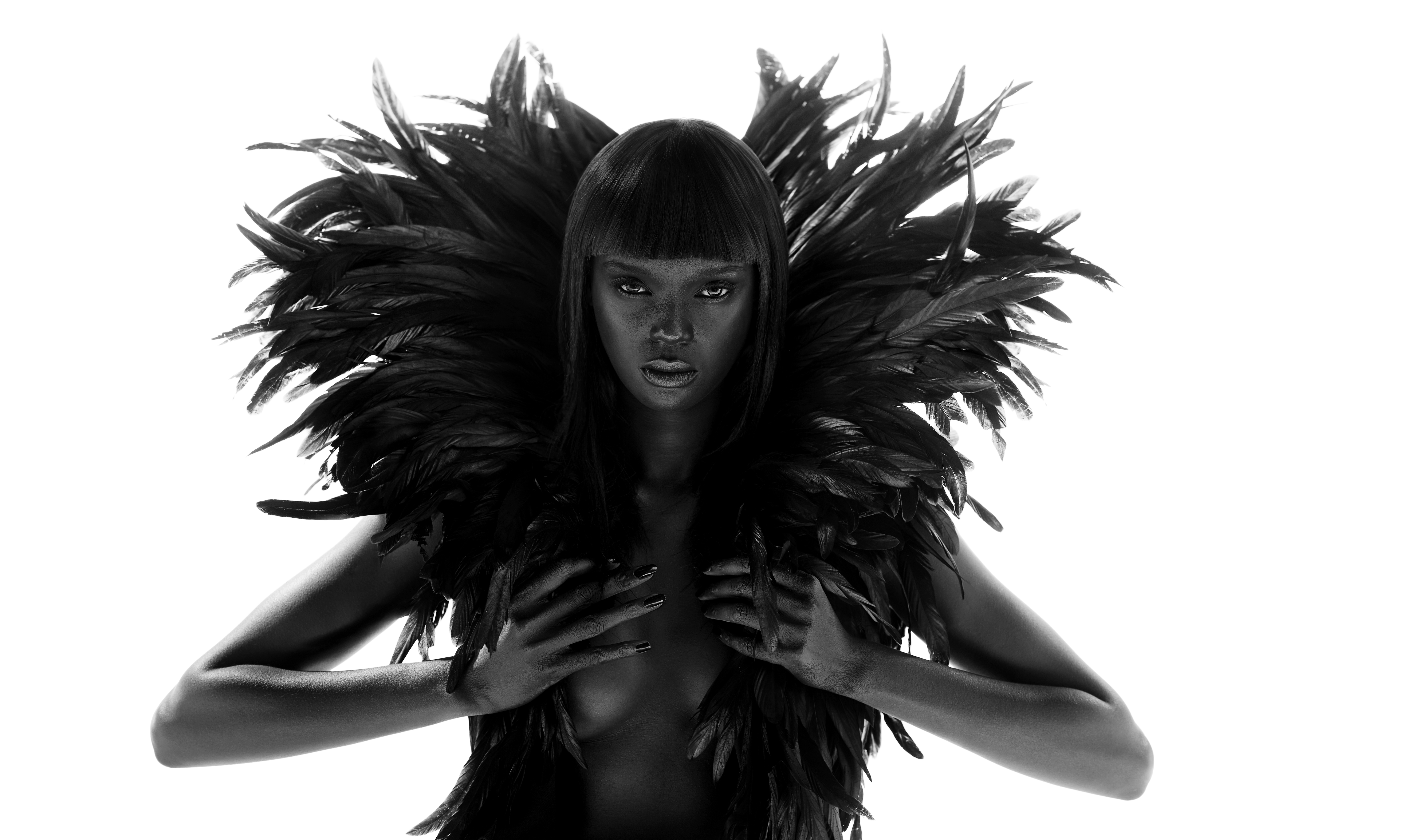 Striking black and white portrait of a dark-skinned adult female with dark hair and feathers, hands posed, set against a stark white backdrop.