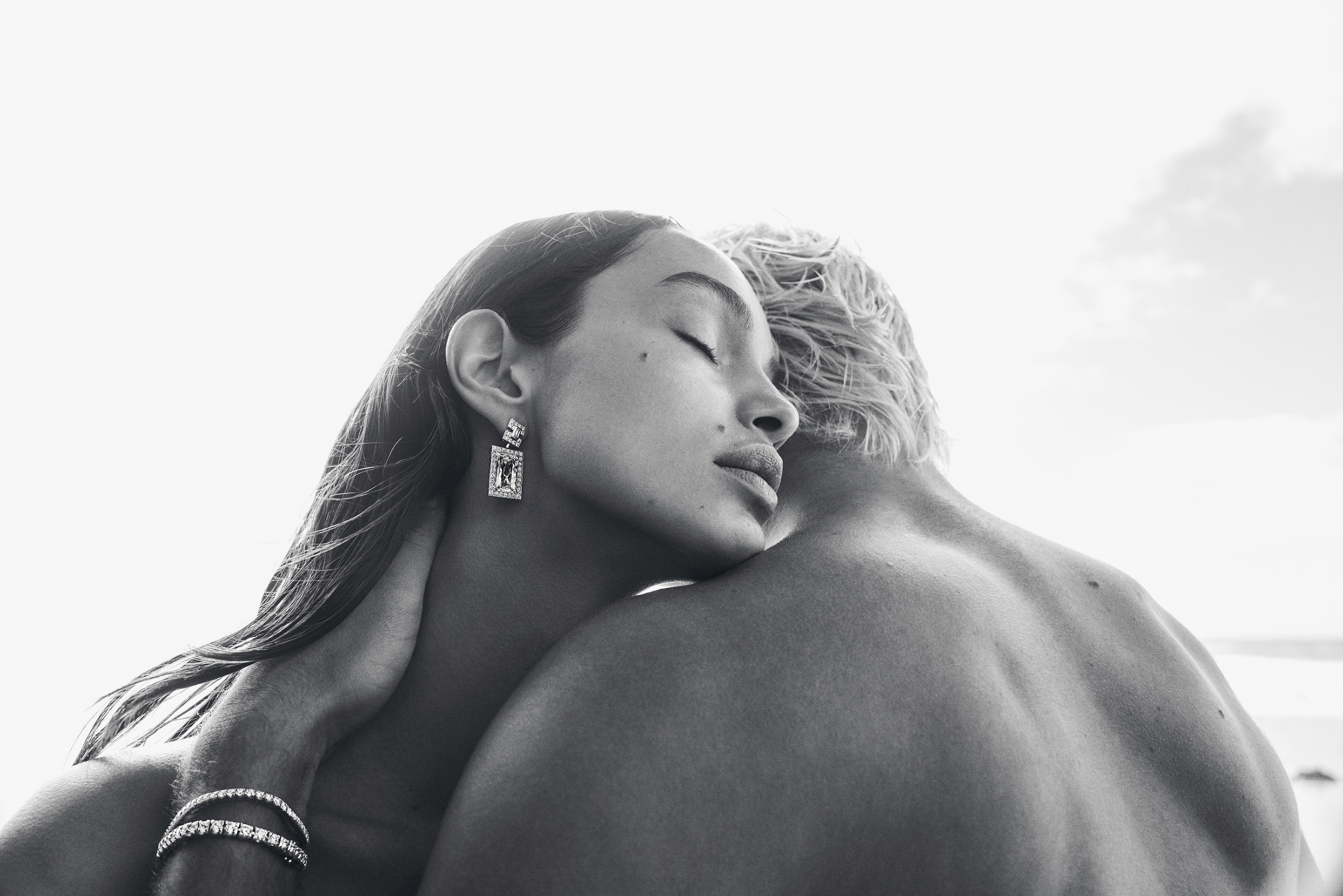Close-up monochrome image of a light-skinned woman with an earring and a bracelet hugging a man against a bright, neutral background.