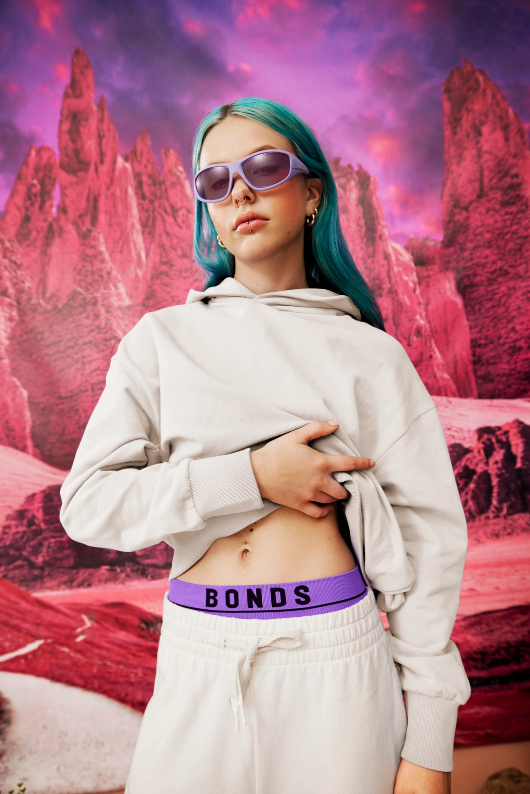 Young woman with bright blue hair wearing purple sunglasses, a cream hoodie, and grey sweatpants against a pink rocky mountain backdrop