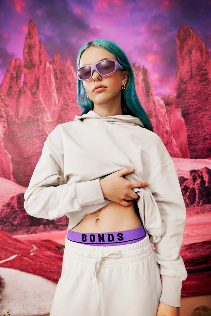 Young woman with bright blue hair wearing purple sunglasses, a cream hoodie, and grey sweatpants against a pink rocky mountain backdrop