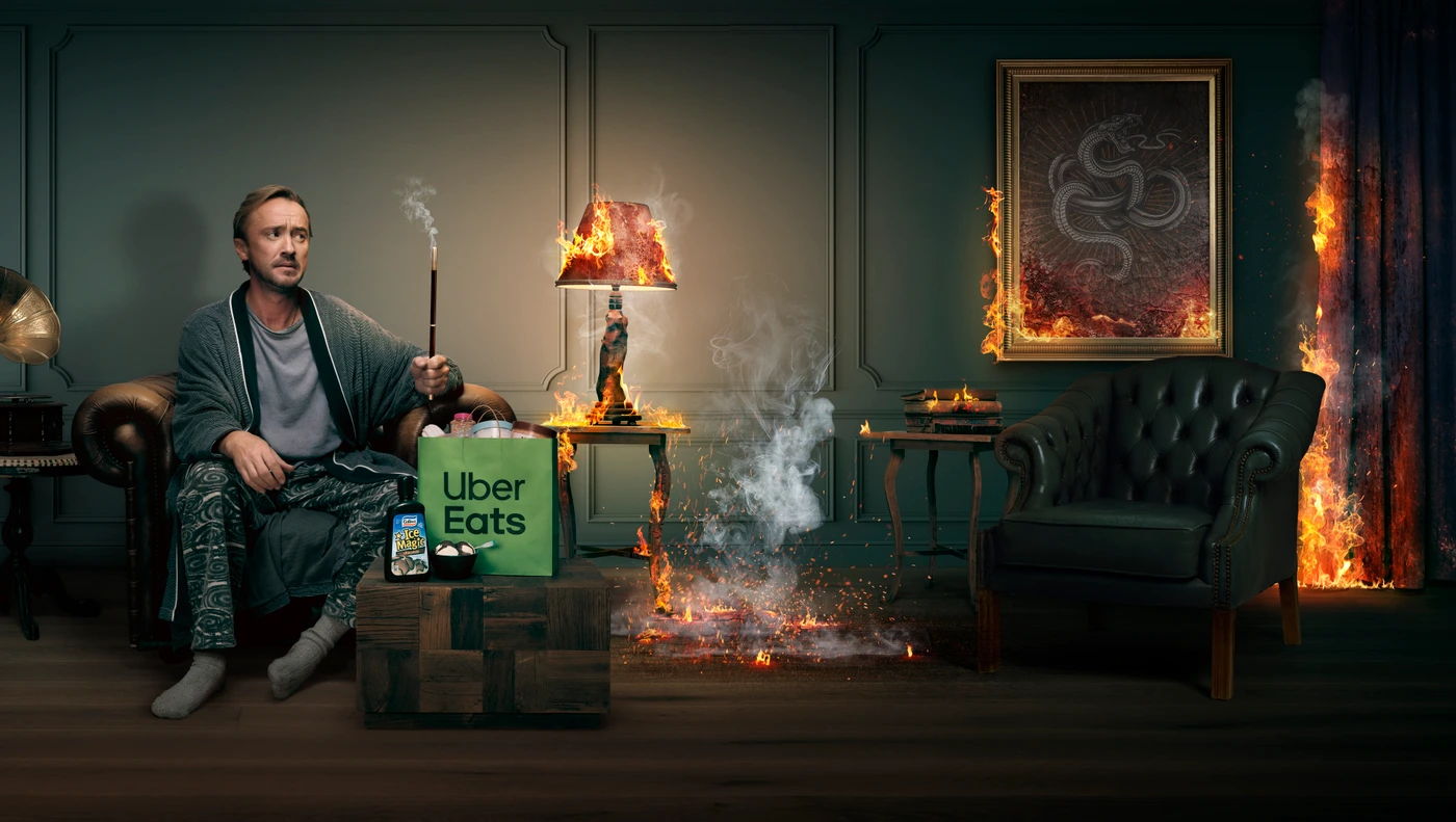 Man holding a wand sitting in a room while a lamp and a painting are on fire beside an Uber Eats bag