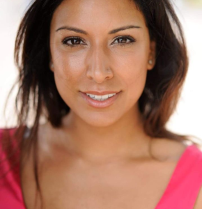 Close-up portrait of a smiling adult woman with brown hair and tan skin wearing a pink top against a blurred neutral background.