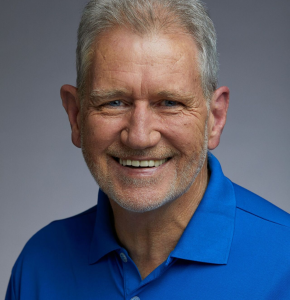Headshot of a mature adult male with light skin and short grey hair wearing a blue collared shirt, posing against a grey backdrop.