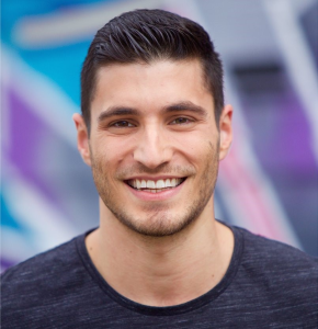 Close-up portrait of a smiling adult male with dark hair, wearing a dark grey shirt, against a blurred, colorful outdoor background.