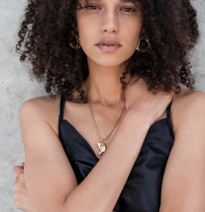 Portrait of a Black woman with curly hair wearing a black top and gold jewelry, posing against a concrete wall in natural light.